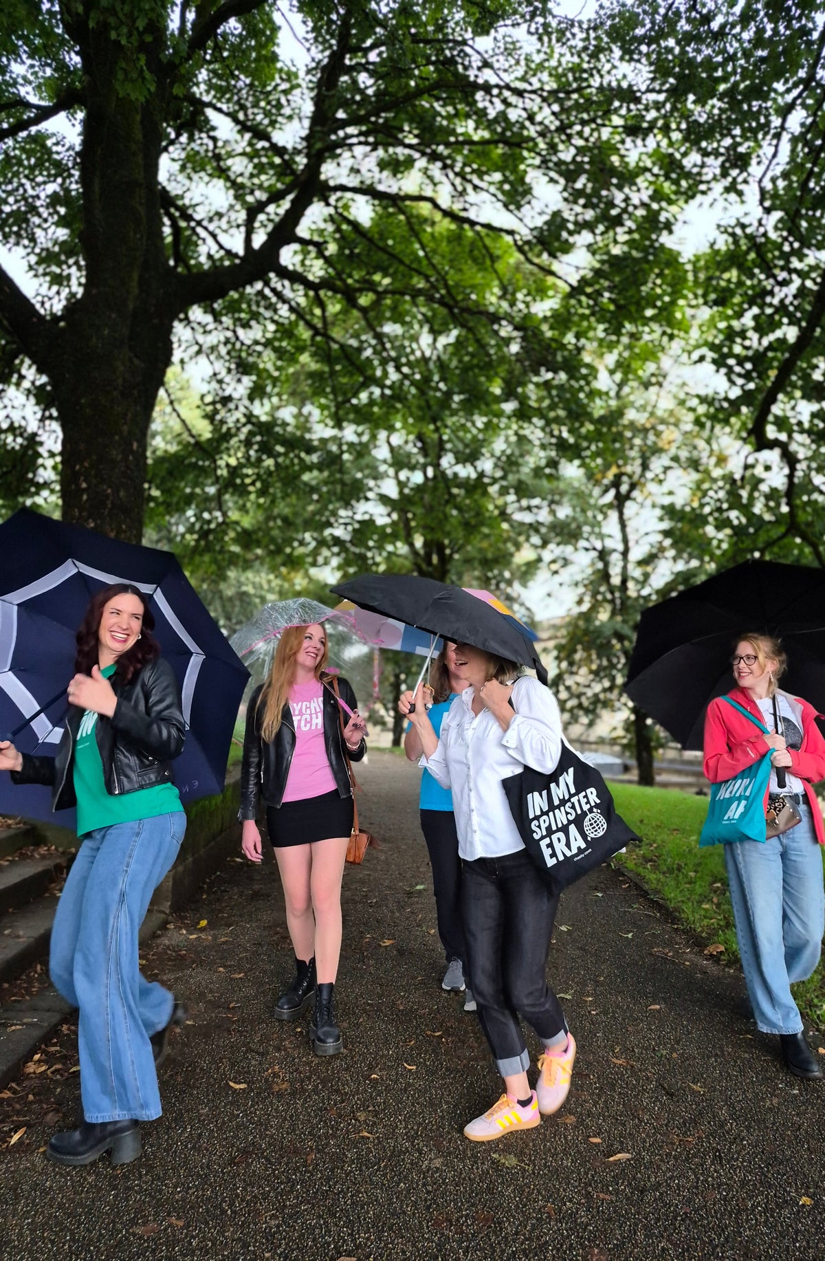 Five women walking on a tree-lined path holding umbrellas, with one person carrying a black tote bag that reads 'IN MY SPINSTER  ERA'.